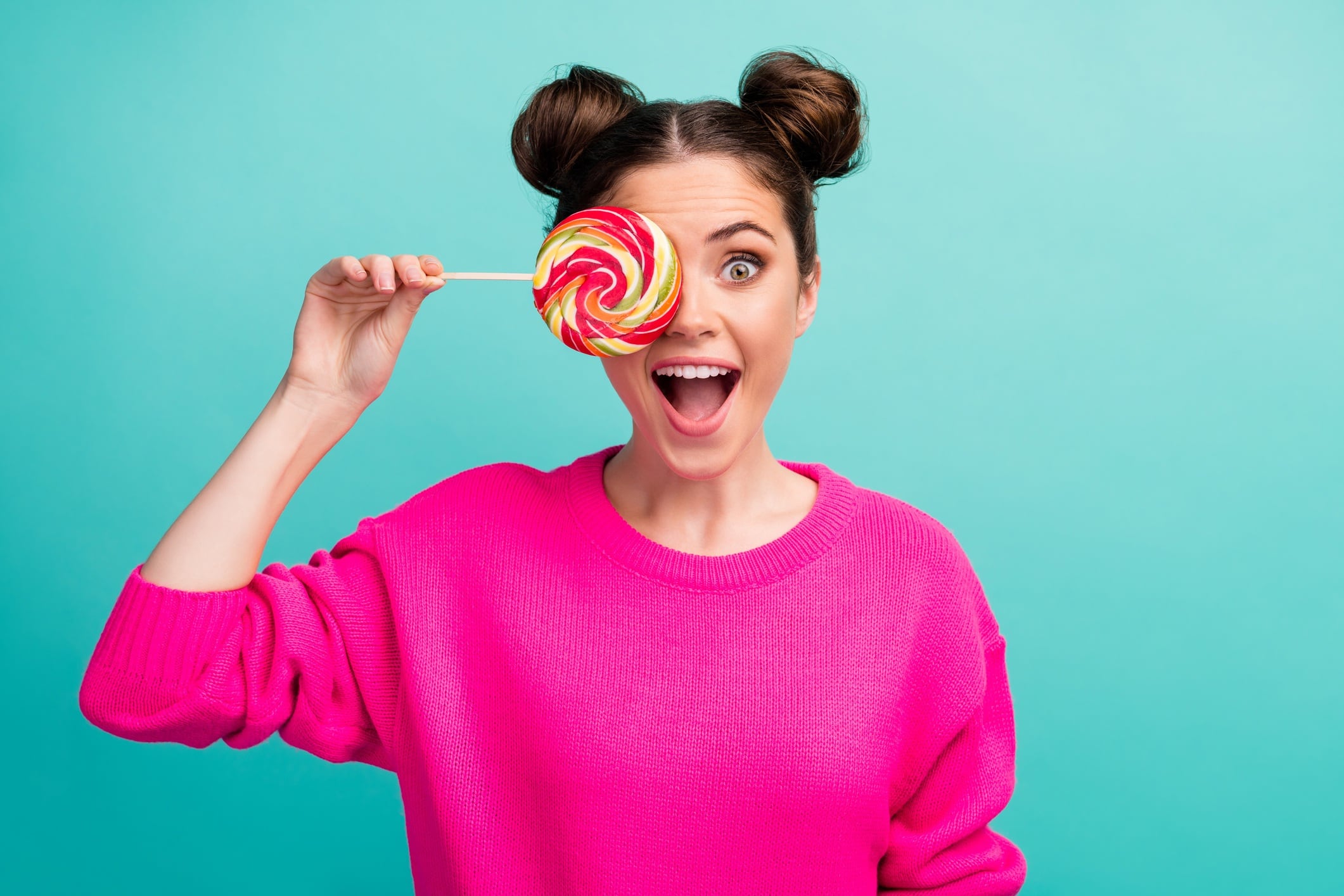 Woman wearing pink jumper and holding lollipop over one eye. Blue-green background.